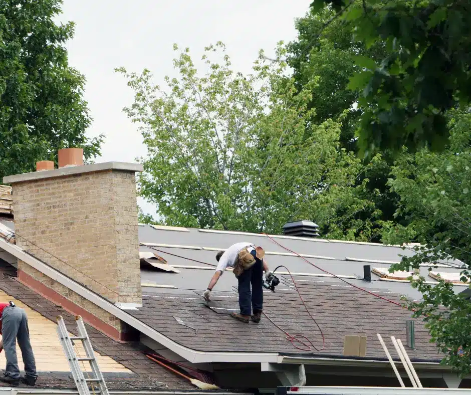 Roofer repairing shingles near a brick chimney surrounded by leafy trees.
