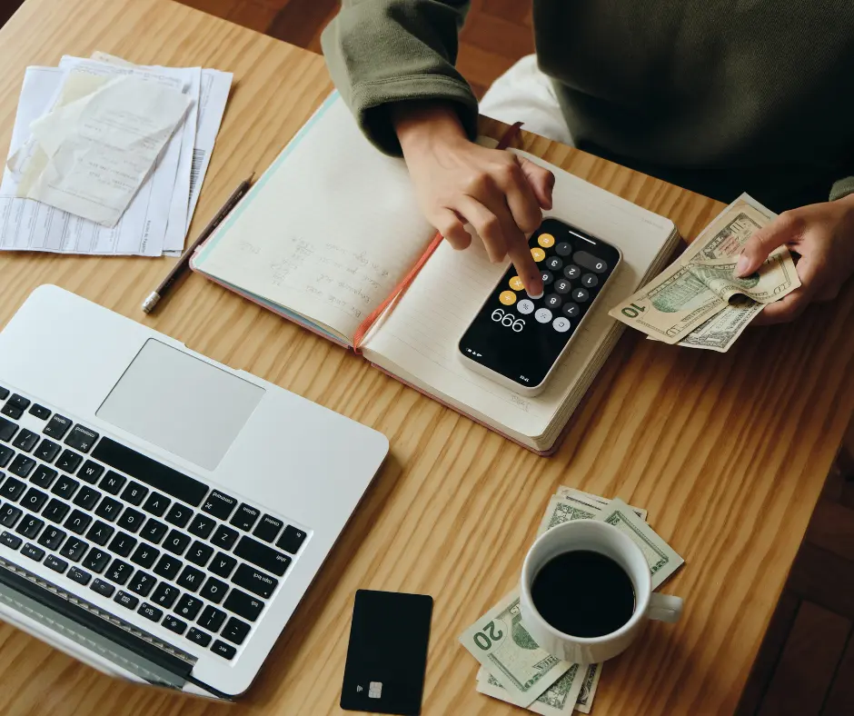 A person calculating finances with a smartphone, dollars, notebook, and laptop on a desk.