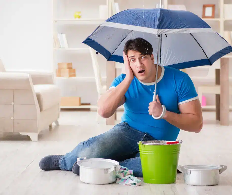 Homeowner sitting under an umbrella catching roof leaks in buckets.